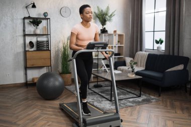 Young attractive sporty african american woman in sportswear having morning workout at modern light apartment. Healthy athletic female jogging while training at treadmill.