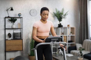 Front view of healthy female athlete having cardio workout indoors. Young healthy african woman training at home while running at treadmill, tapping on control panel screen of machine.
