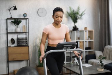 Front view of healthy female athlete having cardio workout indoors. Young healthy african woman training at home while running at treadmill, tapping on control panel screen of machine.