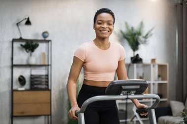 Smiling african american sporty woman in pink t-shirt leaned her hands on treadmill while working out in morning at living room at home, looking at camera.