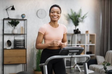 Smiling african american sporty woman in pink t-shirt leaned her hands on treadmill while working out in morning at living room at home, looking at camera.