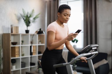 Portrait of active african woman in sportswear using smartphone, running using treadmill at home at morning. Coronavirus Covid 19 social distance. Home workout, stay at home, remote leisure concept.