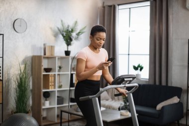 Portrait of active african woman in sportswear using smartphone, running using treadmill at home at morning. Coronavirus Covid 19 social distance. Home workout, stay at home, remote leisure concept.