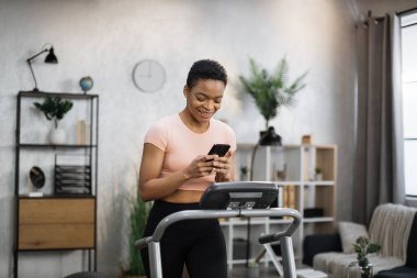 Portrait of beautiful healthy woman working out at home, running on treadmill and using phone for monitoring heart rate and time of training on background of light apartment.