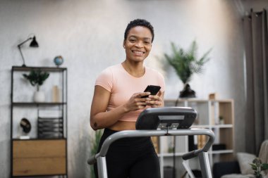 Close up view of young sporty african woman using running machine simulator while making selfie or having video call. Healthy sportswoman having workout on treadmill indoors her home.