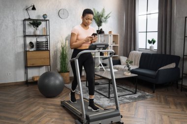 Side view of sporty fit female in pink t-shirt tapping on screen of smartphone, writing message while working out on treadmill, having cardio training at home.