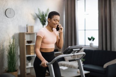 Portrait of young african sportive woman in sportswear talking on smartphone while training at home, doing cardio exercise on treadmill. Concept of sport, health care, communication.