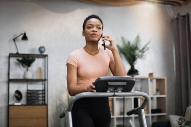 Portrait of young african sportive woman in sportswear talking on smartphone while training at home, doing cardio exercise on treadmill. Concept of sport, health care, communication.