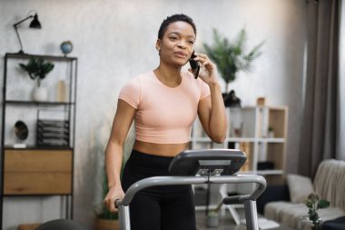 Portrait of young african sportive woman in sportswear talking on smartphone while training at home, doing cardio exercise on treadmill. Concept of sport, health care, communication.