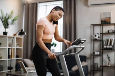 Side view of african sporty woman in pink t-shirt working out in treadmill while having phone call with her relatives or friends. Home activity, lifestyle concept.