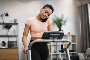 Front view of african sporty woman in pink t-shirt working out in treadmill while having phone call with her relatives or friends. Home activity, lifestyle concept.