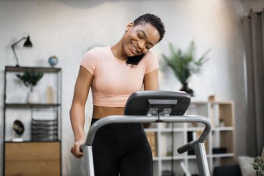 Front view of african sporty woman in pink t-shirt working out in treadmill while having phone call with her relatives or friends. Home activity, lifestyle concept.