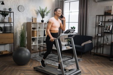 Side view of sporty fit businesswoman in pink t-shirt having video call with business partners using smartphone while working out on treadmill, having cardio training at home.