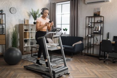 Pozitive african woman in sports clothes and sneakers listening music in headphones while using smartphone, doing running exercise on electric treadmill at morning in fitness room at home.