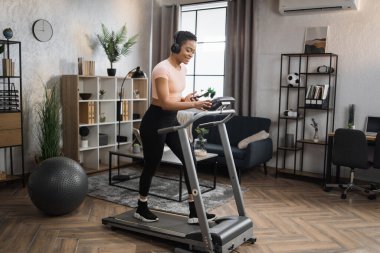 Pozitive african woman in sports clothes and sneakers listening music in headphones while using smartphone, doing running exercise on electric treadmill at morning in fitness room at home.