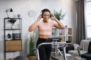 Portrait of young active sporty african american sportive woman in sportswear and headphones training at home, doing cardio exercise on treadmill. Concept of sport, health care.