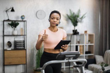 Portrait of active and dynamic young pretty businesswoman with headset and tablet doing sport fitness at home running on treadmill indoor at night showing thumb up. Tone your body. Perfect shape.