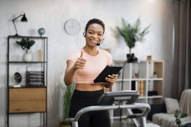 Portrait of active and dynamic young pretty businesswoman with headset and tablet doing sport fitness at home running on treadmill indoor at night showing thumb up. Tone your body. Perfect shape.