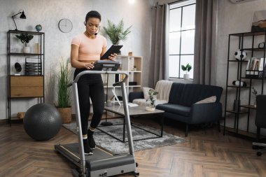 Side view of african woman in sport clothes using headset working on tablet computer while doing cardio training on treadmill at morning at home gym. Concept of sport, health care, business.