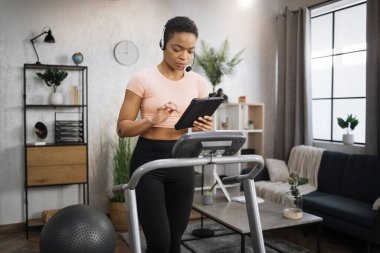 Confident muscular sporty businesswoman in headset working remote from home using digital tablet, while having cardio workout, running on treadmill.