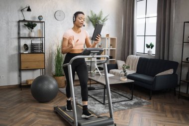 Full length photo of young active african business woman in sportswear and headset training at home working with tablet, doing cardio exercise on treadmill. Concept of sport, business.