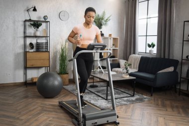 Beautiful african woman in sports clothes and headphones holding bottle of water doing cardio training on treadmill at home gym. Concept of sport, health care, action, remote leisure.
