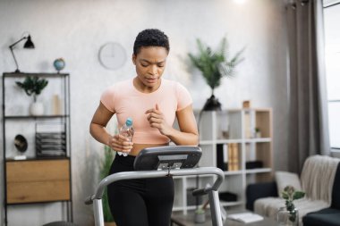 Attractive young sports african american woman holding bottle with water working out, running, doing cardio training on treadmill, indoor on background of modern apartment.