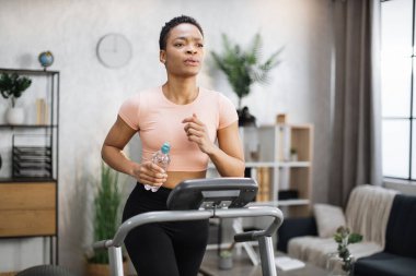 Attractive young sports african american woman holding bottle with water working out, running, doing cardio training on treadmill, indoor on background of modern apartment.