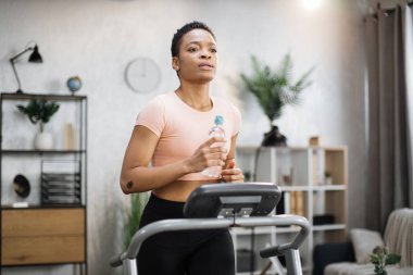 Attractive young sports african american woman holding bottle with water working out, running, doing cardio training on treadmill, indoor on background of modern apartment.