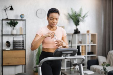 Portrait of active sportive african woman in sportswear opening water bottle while training at home, walking, doing cardio exercise, on treadmill. Concept of sport, health care, thirst.