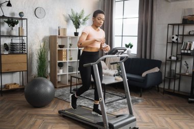 Full length photo of active sportive african woman in sportswear opening water bottle while training at home, walking, doing cardio exercise, on treadmill. Concept of sport, health care, thirst.