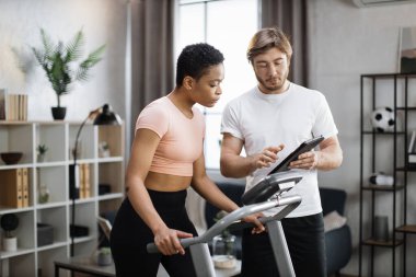 Fitness caucasian man trainer using tablet to show online lesson for african american woman training on treadmill at modern home. Two sporty people engaged in remote workout .