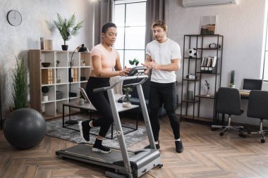 Portrait of focused young african female wearing sportswear and using treadmill and her trainer bearded caucasian male. Home fitness workout sporty woman and man training on running track.
