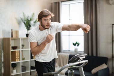 Young attractive sporty man in white t-shirt doing boxing punches while having morning workout at modern light apartment. Muscular strong guy jogging while training at treadmill.