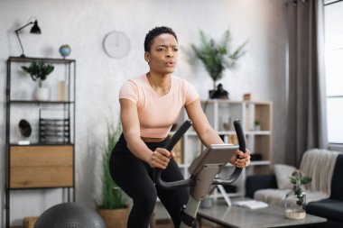 Portrait of focused young african female athlete wearing sportswear and using exercise bike. Home fitness workout sporty woman training on smart stationary bike indoors.