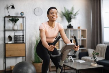 Smiling beautiful african american sports woman in sportswear cycling bike at home on background of light living room. Cardio training, exercising legs, cardio workout indoors.