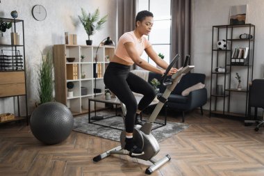 African smiling young sportswoman doing cardio on stationary bike. Attractive female working out on fitness in the morning at home. Woman training on exercise bike indoors.