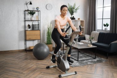 Front view of healthy female athlete having cardio workout indoors. Young healthy african woman training on stationary bike on the background of modern apartment.