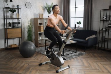 Full length portrait of young sportswoman training at home gym doing cardio on stationary bike. Cardio training, exercising legs, cardio workout indoors.