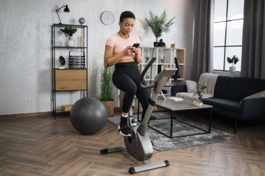 Concentrated african sports women in sportswear tapping on her mobile phone or watching online class, cycling bike at home. Cardio training, exercising legs, cardio workout indoors.