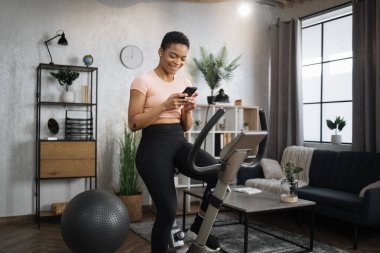 Portrait of young african female wearing sportswear writing message on her smartphone while working out using exercise bike. Home fitness workout sporty woman training on smart stationary bike indoors