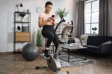 Full length portrait of smiling young african female athlete wearing sportswear and using exercise bike using smartphone. Home fitness workout sporty woman training on smart stationary bike indoors.