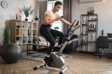 Low angle view of smiling beautiful african american sports woman in sportswear writing message on social media, networks while cycling bike on background of light living room.