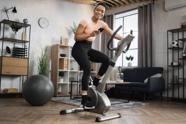 Low angle view of smiling beautiful african american sports woman in sportswear writing message on social media, networks while cycling bike looking at camera on background of light living room.