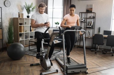 Portrait of focused young caucasian male and african female wearing sportswear using exercise bike and treadmill with phones looking online class how to lose weight.