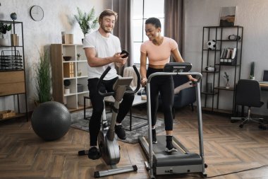 Happy smiling couple doing cardio on stationary bike and treadmill using smart phone to watch online video lessons how to sole weight. Attractive female and male working out on fitness at home.