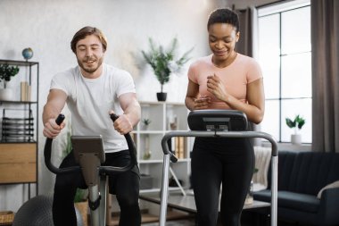 Home fitness workout sporty people training on exercise machines indoors. Portrait of focused caucasian male and african female wearing sportswear using exercise bike and treadmill.