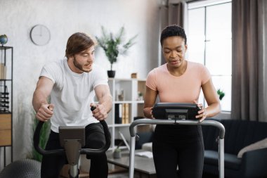 Home fitness workout sporty people training on exercise machines indoors. Portrait of focused caucasian male and african female wearing sportswear using exercise bike and treadmill.