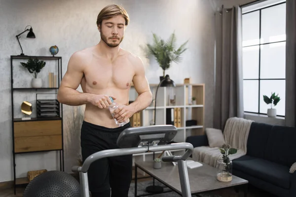 Portrait of active sportive man with muscular bare torso in sportswear opening water bottle training at home, walking, doing cardio exercise, on treadmill. Concept of sport, health care, thirst.