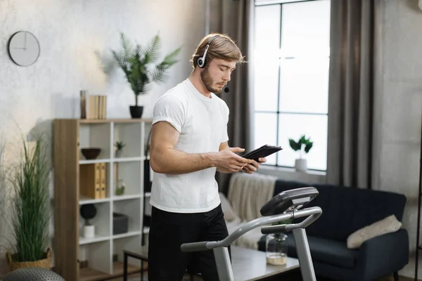 Side view portrait of young active bearded caucasian businessman in sportswear and headset training at home working with tablet, doing cardio exercise on treadmill at evening time.
